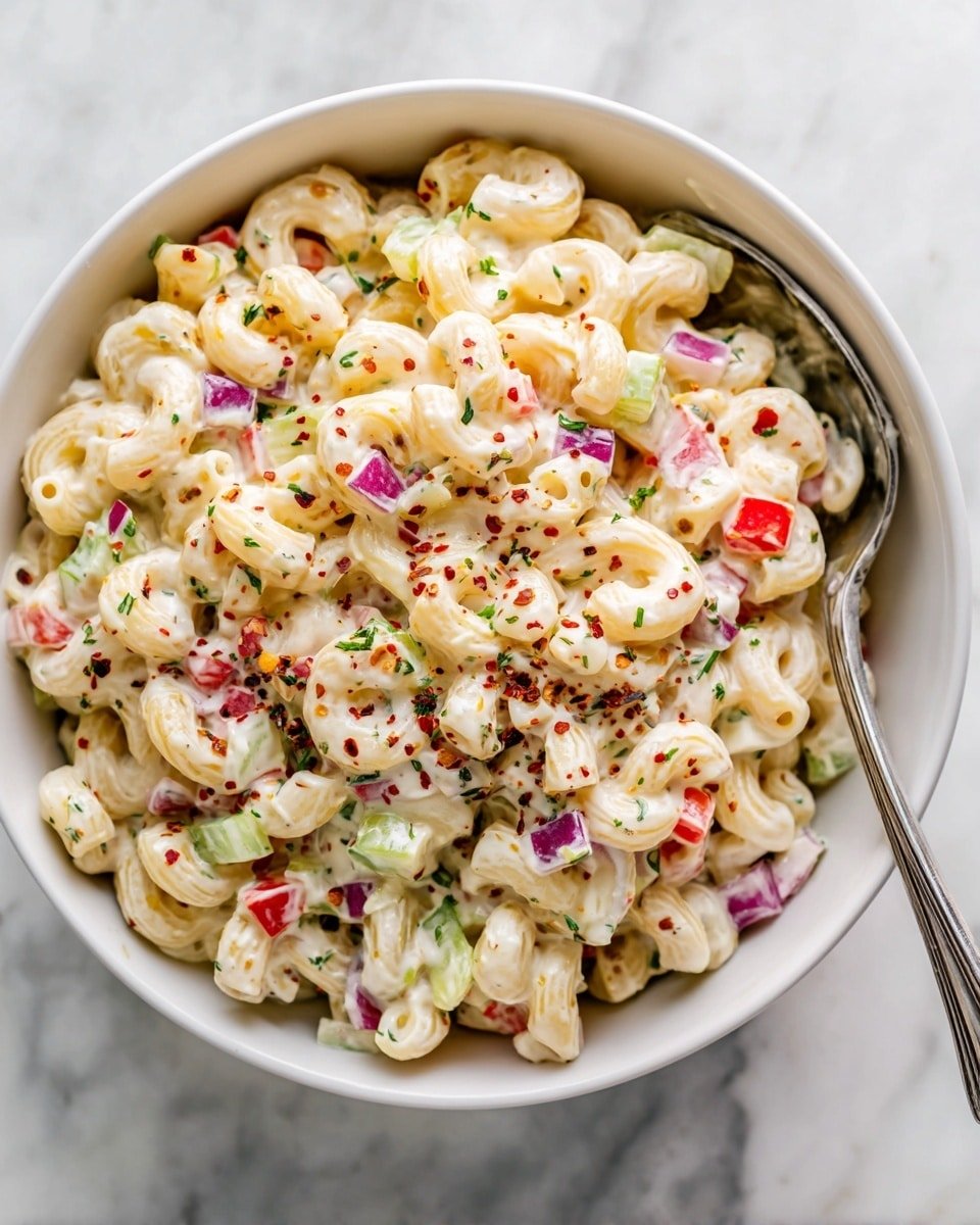 A white bowl filled with creamy macaroni salad showing two main layers: the pasta layer with elbow macaroni coated in smooth, white dressing, and mixed into it are small pieces of bright red bell peppers, pale green celery, and small purple onion chunks. The salad has red pepper flakes and green herbs sprinkled lightly on top, adding specks of red and green over the pale salad. A metal spoon is tucked on the side of the bowl, and the bowl sits on a white marbled surface. Photo taken with an iphone --ar 4:5 --v 7 — Gordon Ramsay Macaroni Salad, Classic pasta salad, creamy mac salad, tangy mustard pasta, easy summer side dish